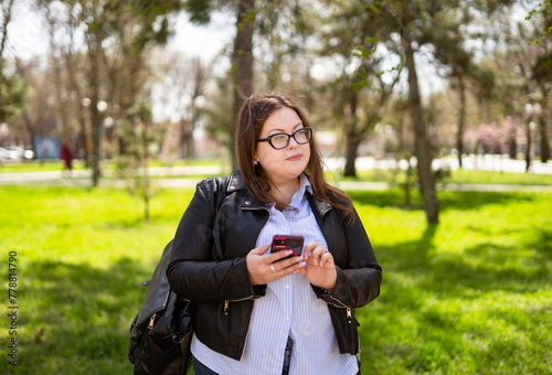 A chubby woman in a short leather bag and casual jeans walks through the park with a phone in her hands. Curvy girl looking at the phone