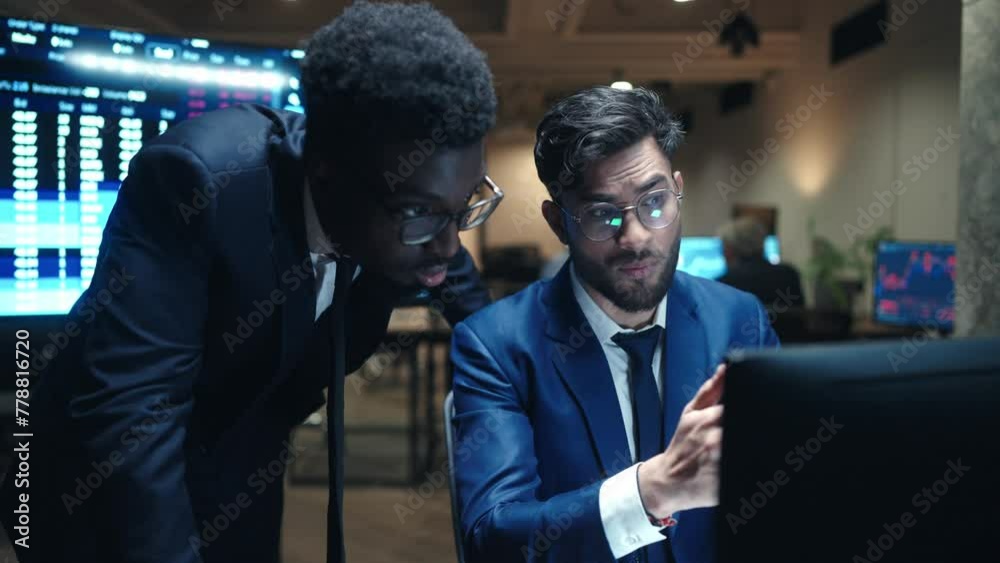 Two multi-ethnic men looking at screen in business office. Hardworking man in blue formal suit explaining something to director. Analysing stock market. On screen visible online data in real time.