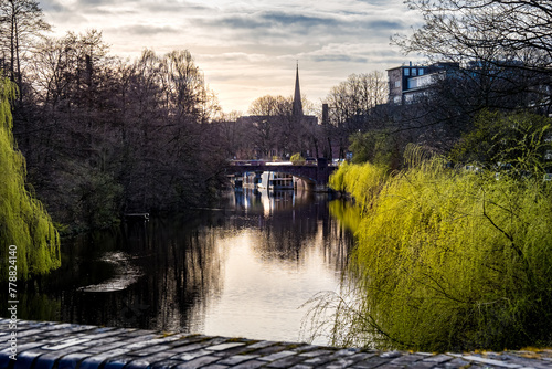 As March evening sets, Wandse river flows through Eilbekkanal canal, flanked by sprouting willows, leading to Wagnerstraßenbrücke bridge, St. Gertrud Church in the background, a serene urban scene.