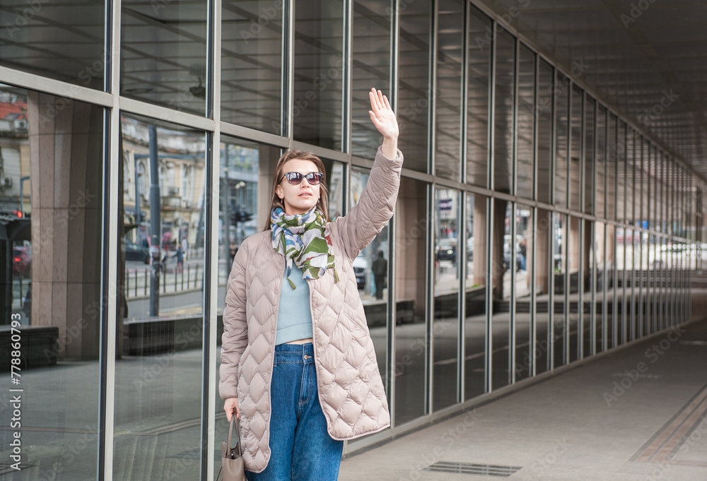 Beautiful stylish woman with sunglasses and bag walking near building with mirror wall on the street and waving her hand hello Beautiful stylish woman with sunglasses and bag walking near building with mirror wall on the street and waving her hand hello