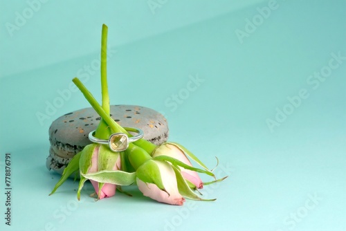 close-up of an engagement silver ring with a gemstone worn on a bouquet of three pink rose buds leaning against a grey speckled macaroni cake on an azure background