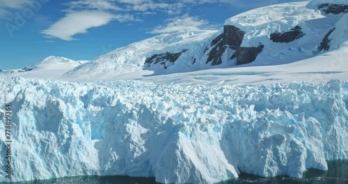 Huge towering ice glacier in Antarctica, snow covered mountain in background, clear blue sunny sky. Ice formation wall cold polar ocean. Aerial arctic winter landscape. Global warming, climate change