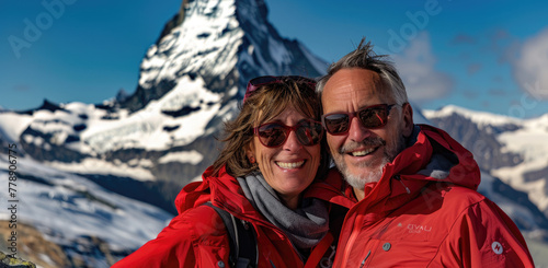 A photo of a middle-aged man and woman in a red jacket, black sunglasses with backpacks smiling at the camera standing on Zermatt mountain with Matterhorn behind them