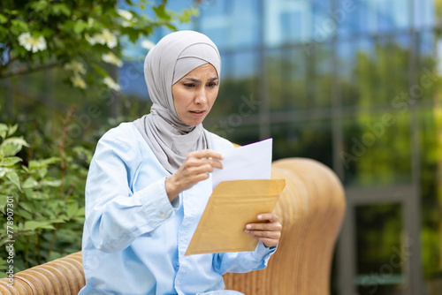 A worried Muslim woman wearing a hijab holding and reading a document or letter while sitting outside on a bench. © Liubomir