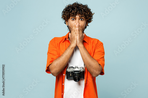Traveler shocked Indian man wear orange casual clothes cover mouth with hand isolated on plain blue background. Tourist travel abroad in free spare time rest getaway. Air flight trip journey concept. photo