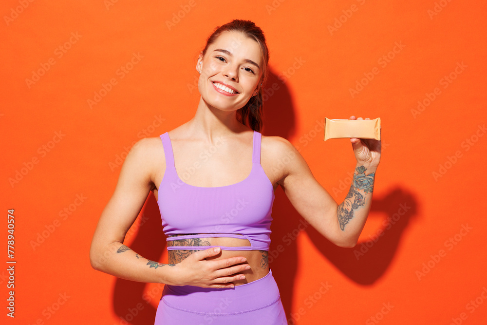 Young fitness trainer instructor sporty woman sportsman wear purple top clothes spend time in home gym protein bar put hand on belly isolated on plain orange background. Workout sport fit abs concept. Young fitness trainer instructor sporty woman sportsman wear purple top clothes spend time in home gym protein bar put hand on belly isolated on plain orange background. Workout sport fit abs concept.