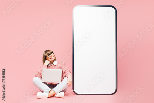 Full body elderly IT woman 50s year old in sweater shirt casual clothes glasses big huge blank screen area mobile cell phone work hold use laptop pc computer isolated on plain pink background studio photo