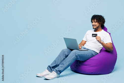 Full body young happy Indian man he wears white t-shirt casual clothes sit in bag chair using laptop pc computer hold credit bank card shopping online booking tour isolated on plain blue background.