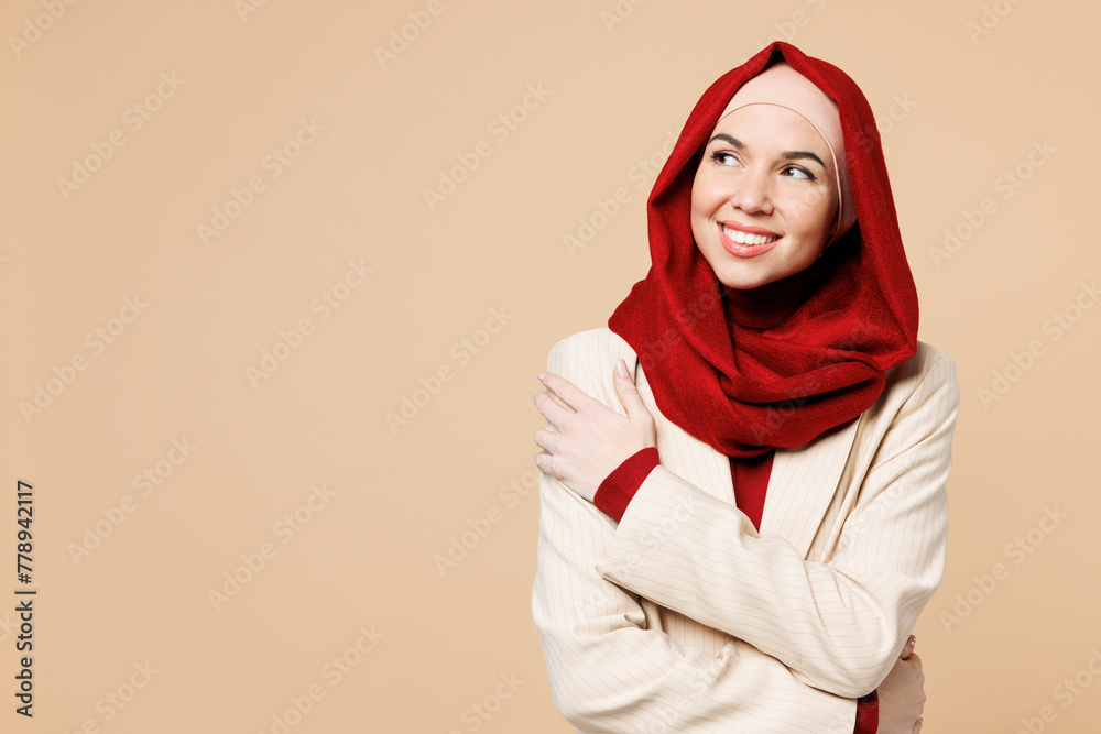 Young smiling Arabian Asian Muslim woman wears red abaya hijab suit clothes hold hands crossed folded look aside isolated on plain beige background studio. UAE middle eastern Islam religious concept. Young smiling Arabian Asian Muslim woman wears red abaya hijab suit clothes hold hands crossed folded look aside isolated on plain beige background studio. UAE middle eastern Islam religious concept.
