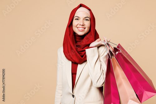 Young Arabian Asian Muslim woman wears red abaya hijab suit clothes shopping hold package bags look camera isolated on plain beige background. Black Friday sale buy day, UAE Islam religious concept.