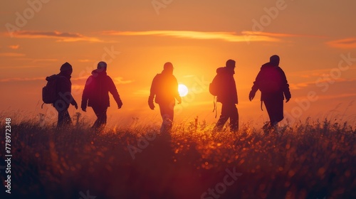 A group of people are walking in a field at sunset. The sun is setting in the background  casting a warm glow over the scene. The people are carrying backpacks