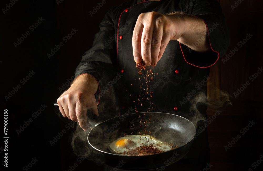 The cook adds aromatic spices to a steaming hot frying pan with fried eggs. Low key concept for a breakfast menu for a hotel with advertising space. Grande cuisine The cook adds aromatic spices to a steaming hot frying pan with fried eggs. Low key concept for a breakfast menu for a hotel with advertising space. Grande cuisine