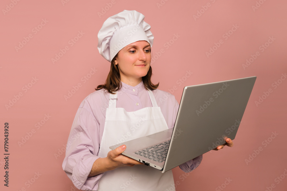 Woman cook with laptop on studio pink background. Portrait of a female person in chef's clothing Woman cook with laptop on studio pink background. Portrait of a female person in chef's clothing