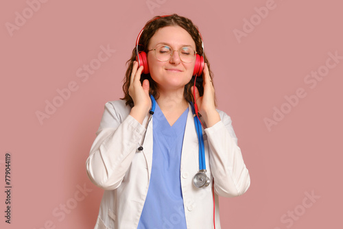 Woman doctor listening to music on headphones, studio pink background. Nurse in uniform with stethoscope on red studio background