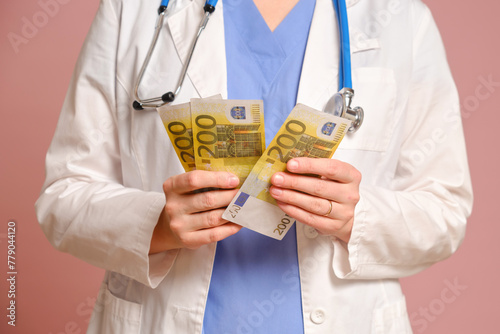 Woman doctor holding money in euro bills in her hands, studio pink background. Nurse in uniform with stethoscope on red studio background © Андрей Журавлев
