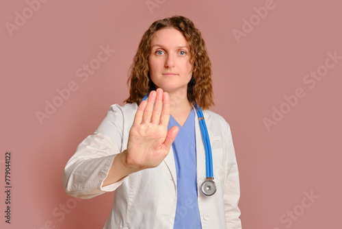 Woman doctor hand gesture stop, studio pink background. Nurse in uniform with stethoscope on red studio background