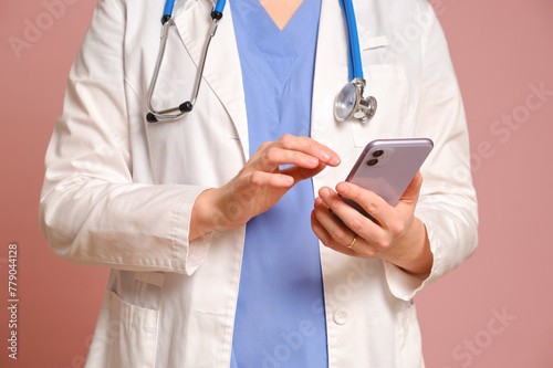 Woman doctor holding a mobile phone in her hands, studio pink background. Nurse in uniform with stethoscope on red studio background