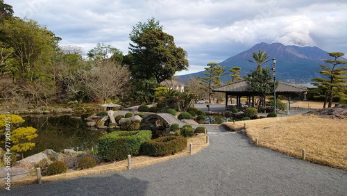 Kagoshima, Japan - 01.29.2020: A trailing leading to a stone bridge over a pond with a pavilion on the side in Sengan-en and Sakurajima at the back releasing smoke under a cloudy sky photo