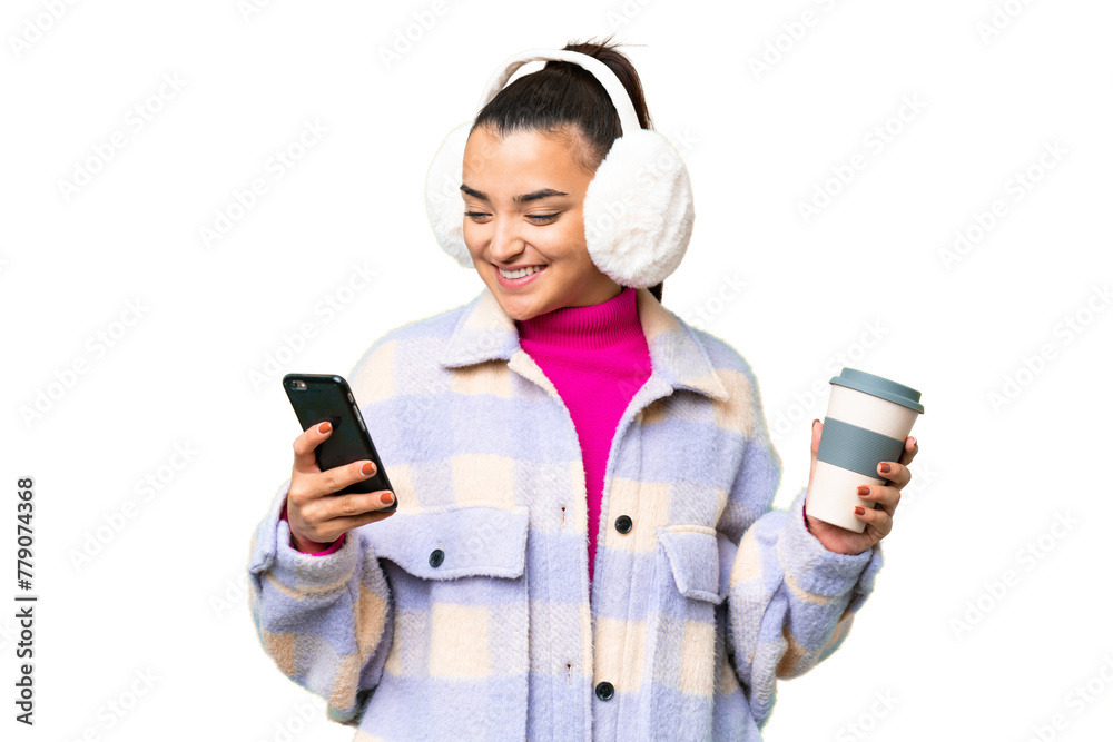 Young woman wearing winter muffs over isolated chroma key background holding coffee to take away and a mobile Young woman wearing winter muffs over isolated chroma key background holding coffee to take away and a mobile