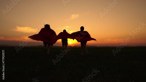 Group of children dressed as superheroes runs across meadow against of setting sun. Silhouette of kids wearing capes fantasizing about superpowers together. Cheerful children create joyful moments