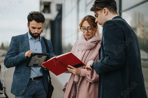 Three young professionals engaging in a business discussion outdoors near a modern urban cityscape, with tablet and paperwork. photo