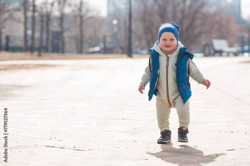 A handsome, cheerful little boy of two years old walks on the street in the spring. A kid in overalls and a blue vest walks through the park. The concept of parenthood, childhood and family.