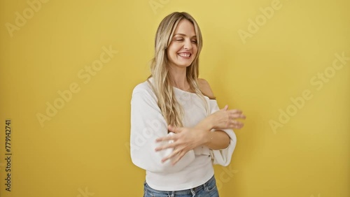 Joyful young blonde woman standing, arms raised in victory, celebrating successful win, eyes closed in happiness over isolated yellow background