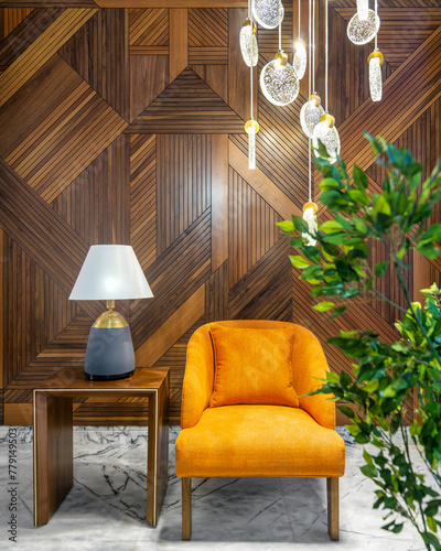 Modern orange armchair, lamp shade on small wooden table, planter with green bushes, and contemporary tall glass chandelier, in a hall with decorated wood cladding wall, and white marble floor