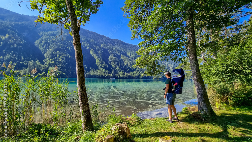 Man with baby carrier looking at scenic view of east bank of alpine lake Weissensee, Gailtal Alps, Carinthia, Austria. Bathing lake surrounded by mountains of Austrian Alps. Untouched nature in summer
