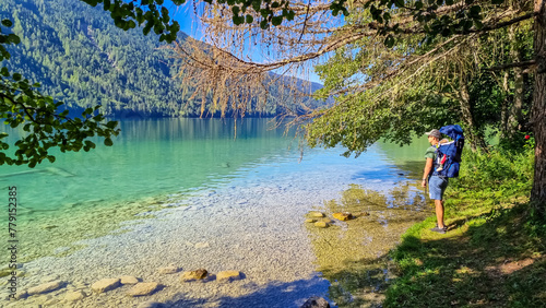 Man with baby carrier looking at scenic view of east bank of alpine lake Weissensee, Gailtal Alps, Carinthia, Austria. Bathing lake surrounded by mountains of Austrian Alps. Untouched nature in summer photo