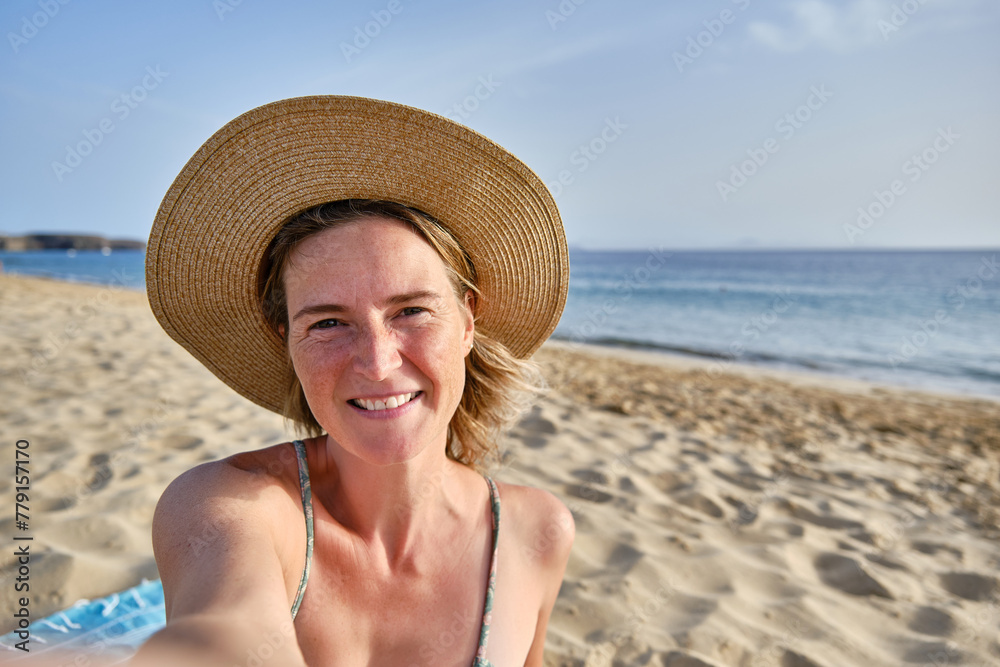 Smiling woman with hat taking a selfie on the beach with the sea in the background