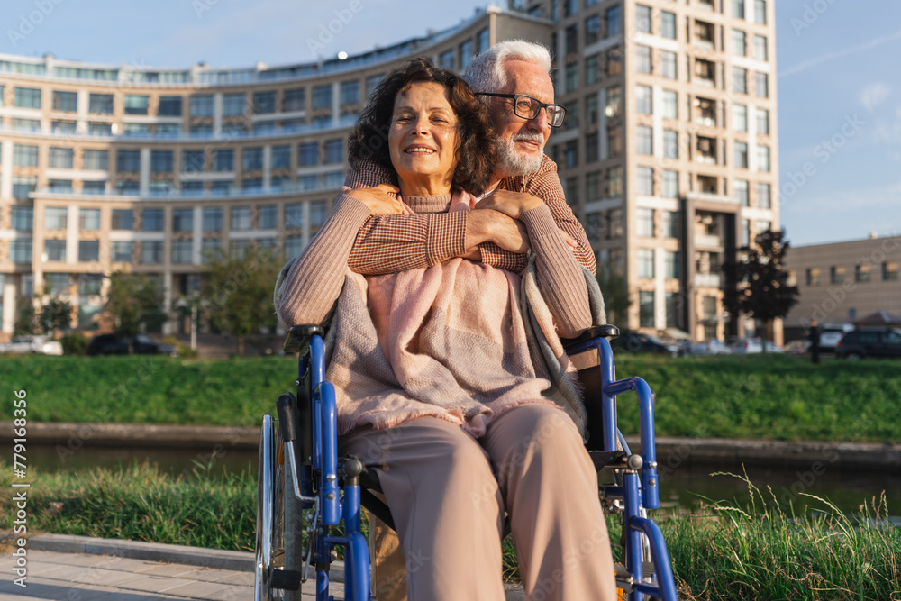 Naklejka premium Senior woman in wheelchair walking with caregiver old man on road in park. Elderly family couple man supporting embracing woman in chair for people with disability outdoor. Rehabilitation