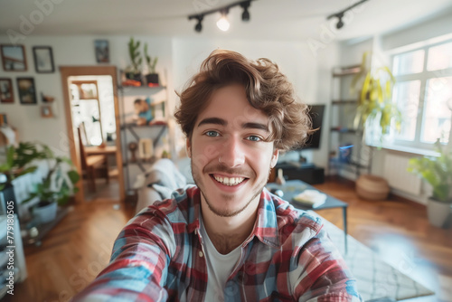 jeune homme entre 25 et 30 ans, avec un anneau dans le nez, les cheveux ondulés mi-longs et les yeux bleus, souriant, qui prend un selfie de lui-même dans son salon à bout de bras photo