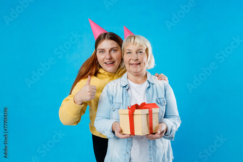 Young daughter and mother together holding birthday present with red ribbon isolated on blue background studio. Human emotions and holiday concept. 30s Caucasian woman giving a thumbs up