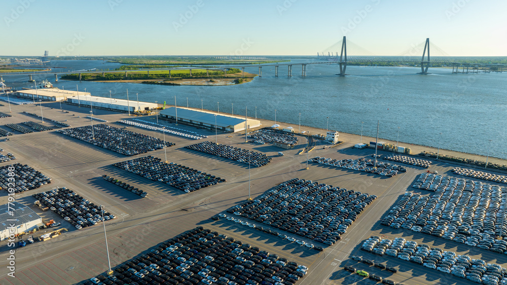 Obraz premium New cars parked in orderly rows at a port parking lot with the expansive Arthur Ravenel Jr. Bridge in the background in Charleston, SC.