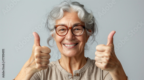 Femme joyeuse levant les pouces en l'air, photographie sur fond blanc, mère de 70 ans avec des lunettes et une chemise beige photo
