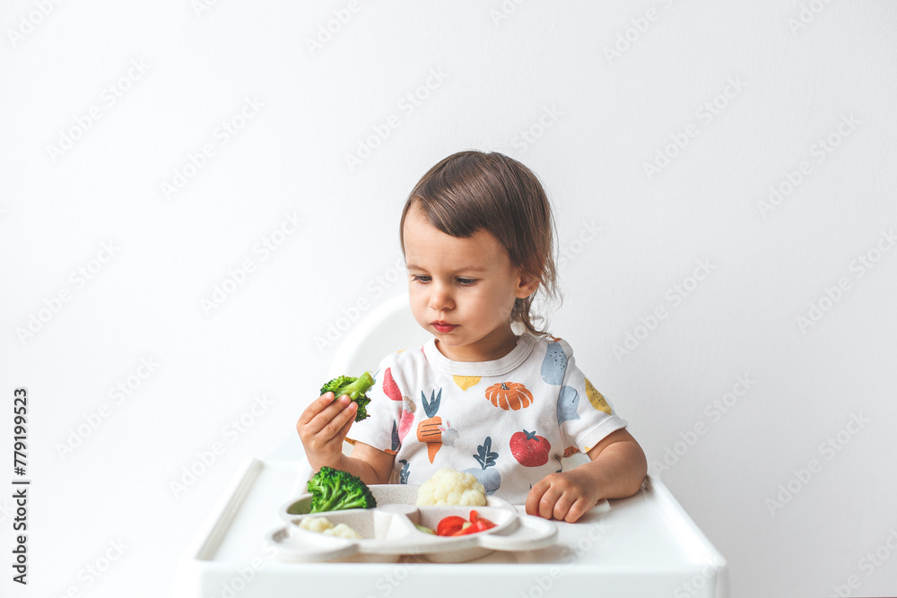 baby 1.5 years old sits in a high chair on a white background eats vegetables, broccoli, cauliflower, cucumbers, tomatoes, concept of children's food, healthy nutrition, vegetarianism