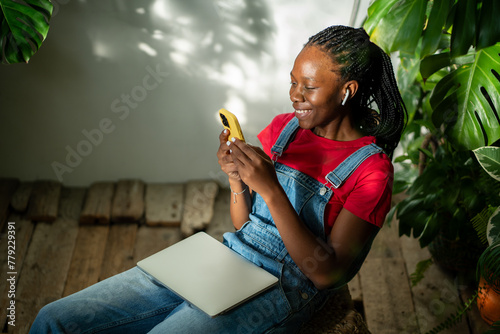 Happy african american woman wearing earphones look screen smartphone listen podcast sit with closed laptop on knees surround by tropical plants. Pleased black girl take break with music on cellphone.