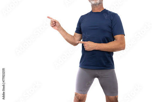 Mature man in sportswear, blue t-shirt and gray shorts, hairy legs, standing and looking forward, pointing with index finger to the blank space on the right. Isolated in a white background.
