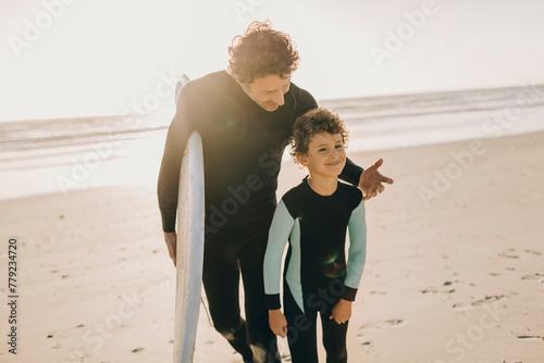 Father and son in wetsuits walking on the beach with a surfboard photo