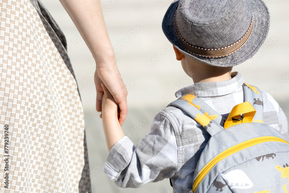 A woman holds a small child by the hand. A little boy in a gray hat and with a backpack on his back, holding his mother's hand. Mom takes the child to school, for a walk, to a children's camp or A woman holds a small child by the hand. A little boy in a gray hat and with a backpack on his back, holding his mother's hand. Mom takes the child to school, for a walk, to a children's camp or
