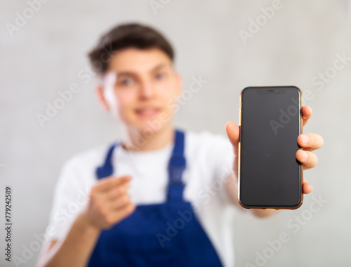 Unfocused handyman in blue jumpsuit shows dark empty mobile phone screen and points at device with hand finger.Device close up, gray background, studio shot photo