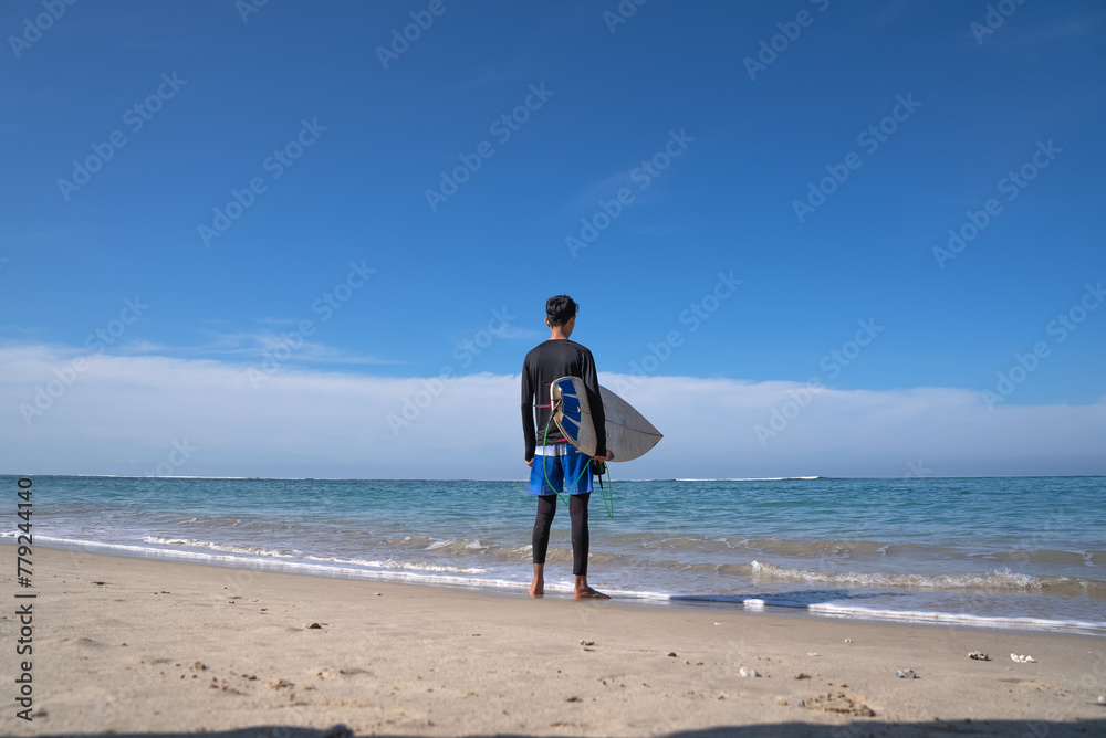 A surfer stands on the beach carrying a surfboard
