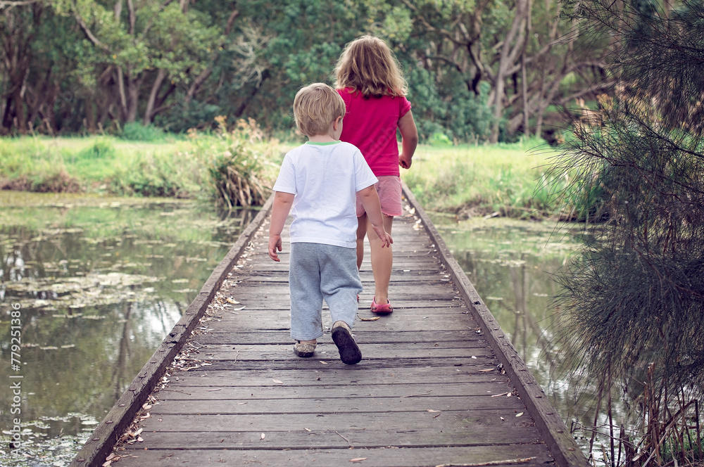 Young children, boy and girl, walking away across timber bridge crossing water, toward a forest of trees Young children, boy and girl, walking away across timber bridge crossing water, toward a forest of trees