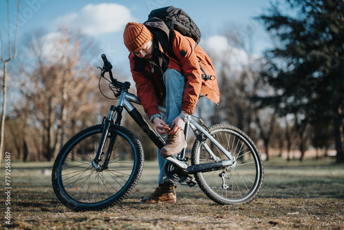 A person wearing a beanie and orange jacket tying their shoe next to a mountain bike in a sunny park setting.