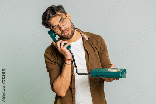 Tired bored Indian man talking on wired vintage telephone of 80s, fooling, making silly faces, exhausted of tedious story, not interested in communication talk. Arabian guy isolated on gray background