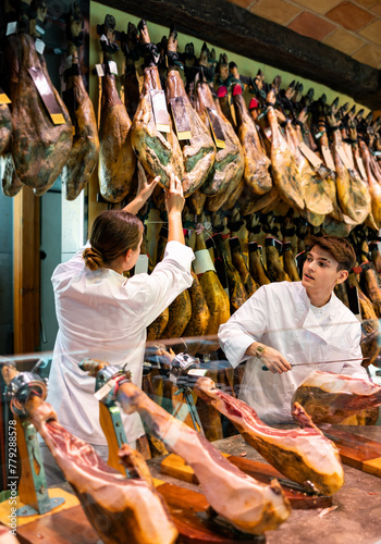 Caucasian young man and woman in uniform carving jamon slices from dry-cured pig's leg in shop. Jamoneria workers cutting ham with knife.