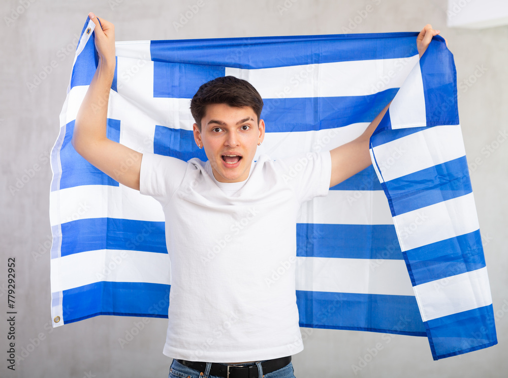 Φωτογραφία Middle-aged positive man in casual clothes holds unfurled flag of Greece in hands raised above head against gray wall, studio shot
