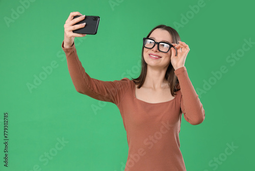 Young woman in eyeglasses taking selfie on green background