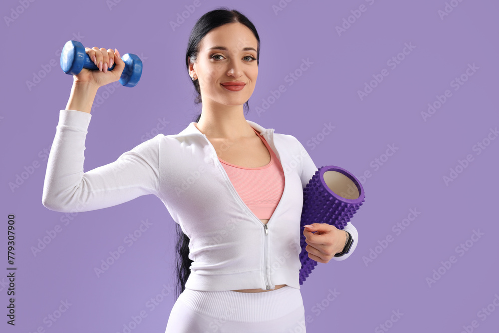 Portrait of sporty young woman with dumbbell and body roller on lilac background - obrazy, fototapety, plakaty 